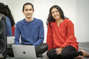 Jay Afrisando, a medium-skin-toned male with his hair bunned, and Siska Aprisia, a medium-skin-toned female with her hair untied, sit side-by-side with their legs folded. They smile. In front of Afrisando is his grey laptop. On the left back, a mirror partly reflects their poses from behind.